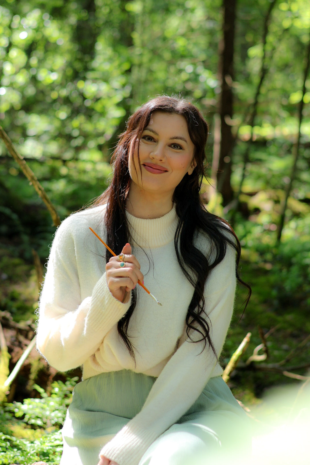 Portrait of Finnish contemporary artist Jenni Inberg smiling in a forest while holding a paintbrush.
